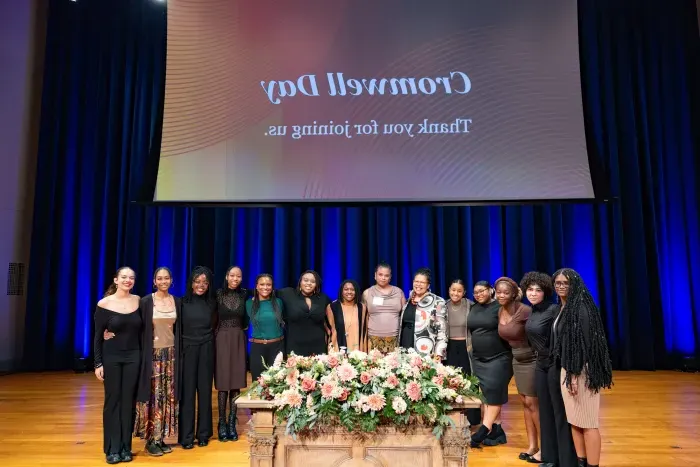 Several students pose for a photo while standing on the stage in front of a large screen that reads, "Cromwell Day. Thank you for joining us." President Sarah Willie-LeBreton poses with them in the center.